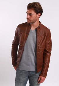 Giacca di pelle marrone con texture liscia, taglio slim, colletto alto e chiusura con zip, indossata sopra un maglione grigio e jeans blu.