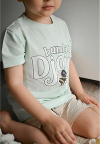 Vaaleanvihreä puuvillapaita, jossa on painettu muotoilu, jossa lukee "humlan Djoy" ja kuvitus mehiläisestä. Lyhyet hihat, rento istuvuus.