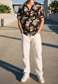 Homme debout à l'extérieur portant une chemise noire à manches courtes à motif floral, un pantalon blanc et des baskets blanches, les mains dans les poches.
