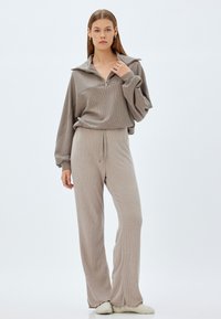 Conjunto de lounge de color beige claro con relieve que incluye una parte superior con cremallera y cuello, y pantalones con cordón y un ajuste holgado, todo con una textura suave.