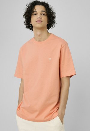 Jeune homme aux cheveux bouclés portant un t-shirt pêche à manches courtes avec une petite broderie blanche de palmier, mains dans les poches, fond neutre.