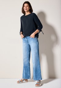 Top lungo a maniche lunghe di colore blu navy con dettagli a fiocco, abbinato a jeans a gamba larga di colore blu chiaro e sandali argentati con cinturino regolabile.