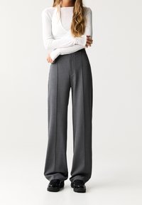 Pantalons larges gris en tissu léger avec des plis, associés à un top blanc à manches longues. Des baskets noires complètent la tenue.