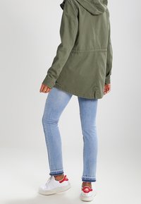 En person som bär ljusblå tajta jeans, vita sneakers med röda detaljer och en olivgrön hoodie, står mot en enfärgad bakgrund.