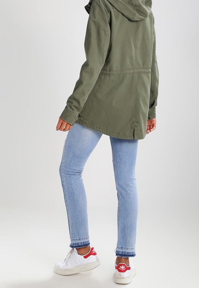 En person som bär ljusblå tajta jeans, vita sneakers med röda detaljer och en olivgrön hoodie, står mot en enfärgad bakgrund.