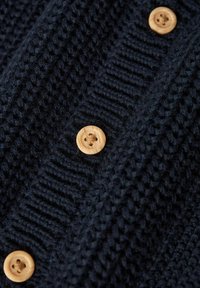 Marineblå strikket sweater i close-up med et struktureret mønster og træknapper med fire huller. Detaljeret ribstrik langs sømmene.