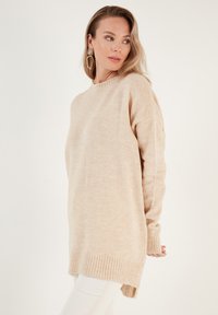 Pull en tricot beige avec un col ras du cou, des manches longues, des poignets côtelés et un ourlet asymétrique. Texture douce et coupe décontractée.