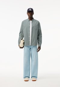 Camisa de cuadros verde y blanca de manga larga, pantalones vaqueros de pierna ancha en azul claro, camiseta blanca, gorra navy con "L", sosteniendo un bolso de crema.