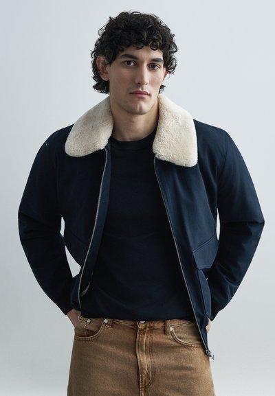 Veste bleu marine avec un col en fausse shearling crème, fermeture éclair à l'avant, poches latérales et texture de tissu lisse. Portée par-dessus une chemise noire.
