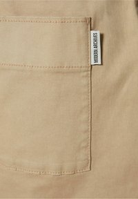 Beige bomullstyg med en lapplomme och en vit etikett som står "MODERN ARCHIVES." Texturen är jämn med synliga stygn.