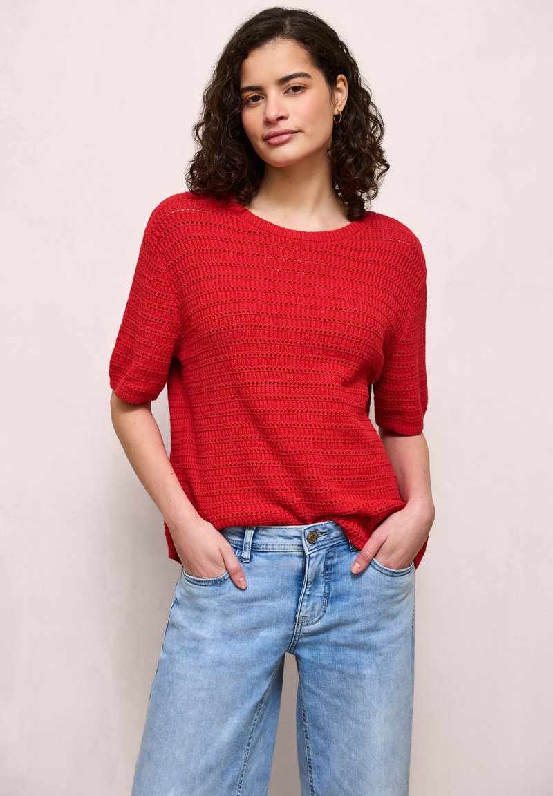 Femme aux cheveux bouclés portant un haut en tricot rouge à manches courtes et un jean bleu clair, les mains dans les poches, sur un fond uni.