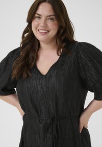 Zwart, gestructureerd jurkje met een V-hals, pofmouwen en een bijpassende strikriem in de taille. Soepele, lichte stof met een subtiele glans.