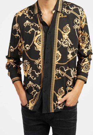 Chemise noire à manches longues avec motifs baroques dorés et motifs de chaînes portée sans être rentrée par une personne les mains dans les poches.