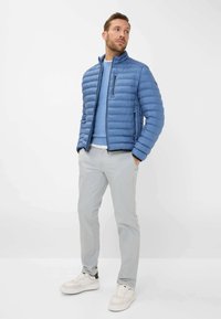 Polsternde blaue Jacke mit hohem Kragen, Reißverschlusstaschen und grauen Kontrast-Hosen, kombiniert mit weißen Turnschuhen, vor einem schlichten Hintergrund präsentiert.