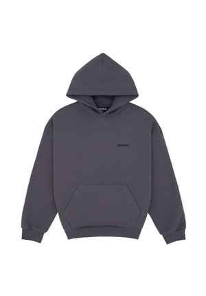 Femme portant un sweat à capuche gris foncé surdimensionné, un pantalon cargo large vert clair avec accessoire chaîne, de grandes lunettes de soleil noires et des chaussures noires pointues.