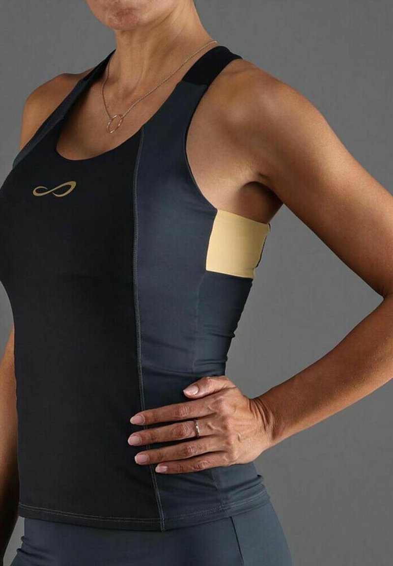 Donna che indossa un top atletico senza maniche aderente nero e grigio con pannello laterale beige, mano poggiata sull'anca, sfondo grigio.