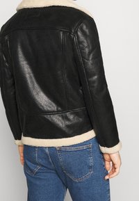 Czarna skórzana kurtka z miękkim, kremowym kołnierzem i mankietami z owczej wełny, fakturowana powierzchnia i dopasowany krój, zestawiona z niebieskimi dżinsami.