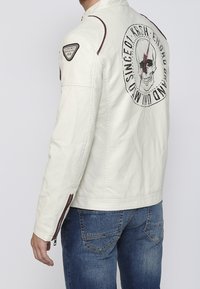 Giacca bianca con grafica di teschio e testo sul retro, dettaglio con cerniera sulla manica, indossata con jeans blu.