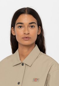 Beige bundový kabát s velkým límcem a logem. Tkanina vypadá hladce s mírným leskem, umístěná na jednoduchém bílém pozadí.