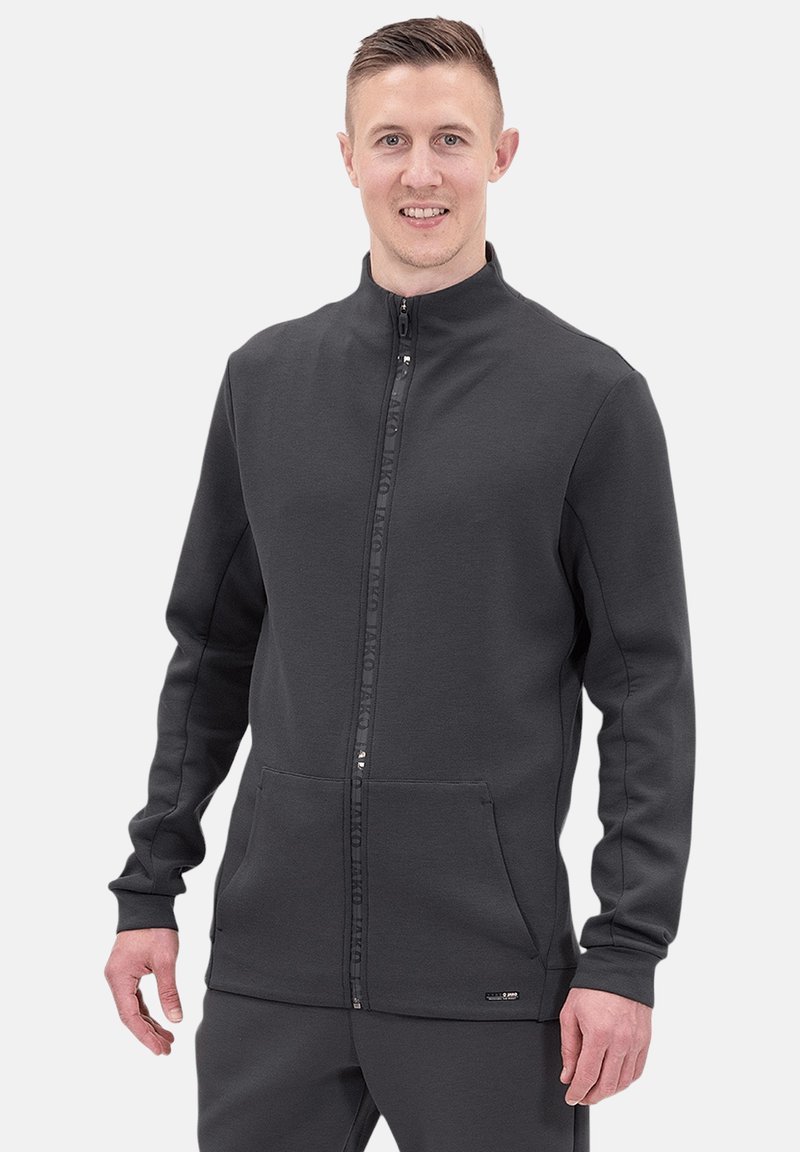 Graue Zip-Jacke aus weichem Material. Hat einen hohen Kragen, Seitentaschen und einen Markenstreifen entlang des Reißverschlusses. Regelmäßige Passform.