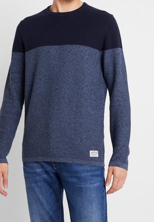 Mann trägt einen langärmligen Pullover in Marineblau und Blau mit Farbblockmuster, kombiniert mit blauen Jeans vor einem einfarbigen Hintergrund.