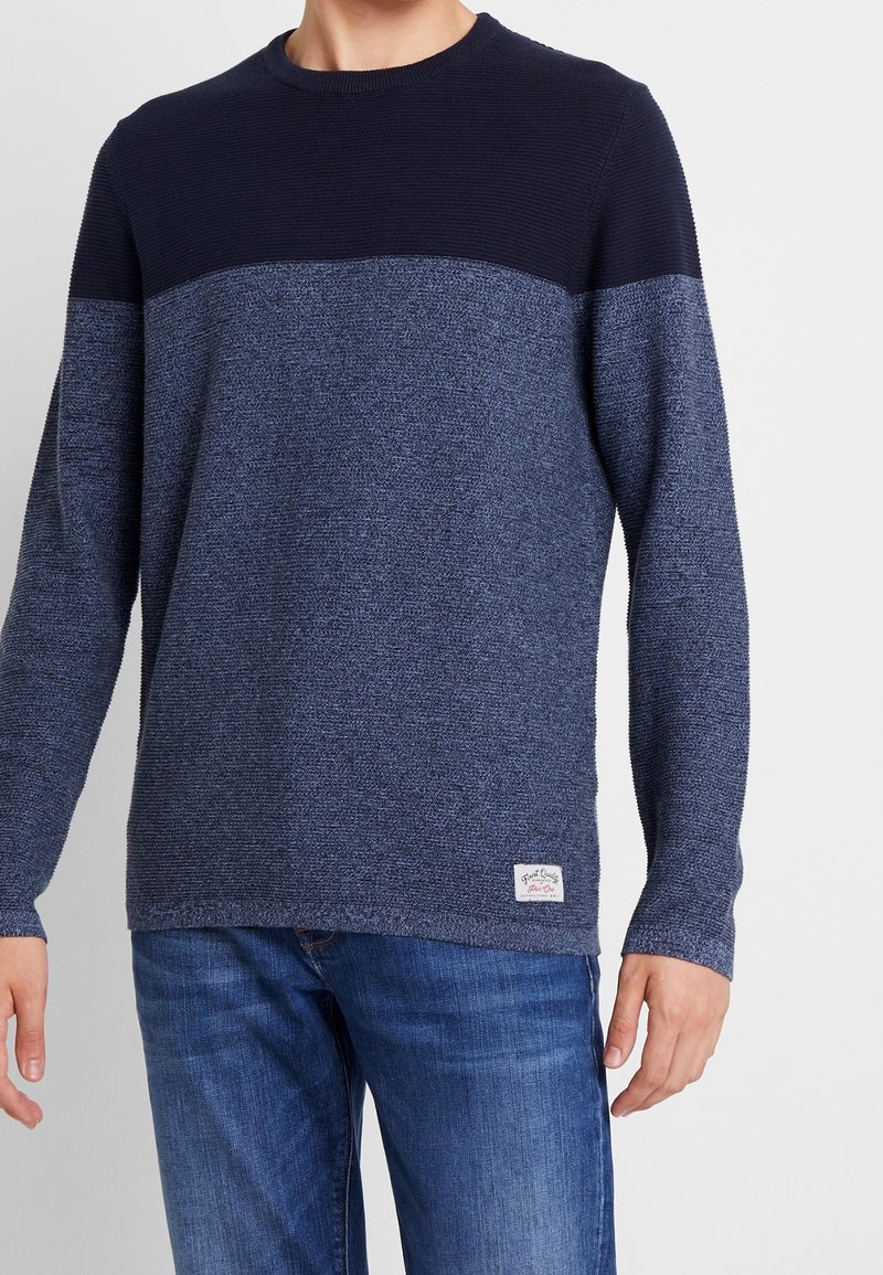 Homme portant un pull à manches longues bicolore bleu marine et bleu, associé à un jean bleu, sur un fond uni.