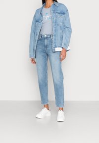 Ljusblå denimjacka över en grå t-shirt med "Calvin Klein Jeans"-logotyp, kombinerat med matchande ljusblå jeans och vita sneakers.