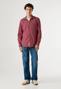 Camicia a quadri rossi e bianchi con colletto in denim, abbinata a jeans blu e sneakers bianche. Il modello si trova di fronte a uno sfondo neutro.