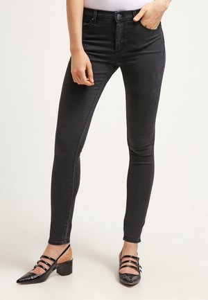 Femme portant un jean skinny noir avec poches avant, associé à des chaussures noires à brides à petit talon, debout avec une main dans la poche.
