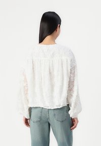 Weiße, florale Spitzenbluse mit lockerer Passform, abfallenden Schultern und langen Ärmeln, kombiniert mit hellblauen, hochbundigen Jeans.