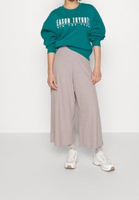 Teal oversized sweatshirt med vit text, parat med ljusbeiga vida byxor. Vita sneakers och mönstrade strumpor synliga.