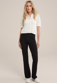 Bílý krátký rukáv pletený top s vlnkovaným lemem a zapínáním na knoflík, spárovaný s černými upnutými kalhotami a bílými teniskami. Jednoduché pozadí.