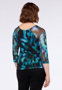 Donna con capelli castani corti, che indossa un top a maniche lunghe blu-verde e nero con effetto tie-dye e pantaloni neri, rivolta di spalle su uno sfondo bianco.