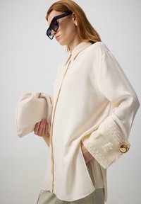 Blouse crème clair avec un col haut, fermeture éclair à l'avant et manches texturées. Notez le détail des boutons décoratifs et le sac à main assorti.