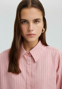 Chemise boutonnée rose clair avec de fines rayures blanches, un col classique et un tissu lisse. Le modèle porte de grandes boucles d'oreilles en or.