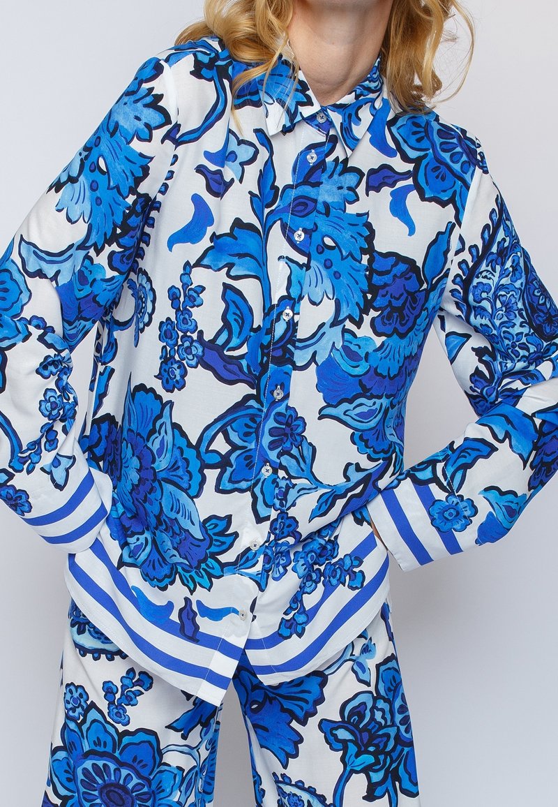 Femme portant une chemise blanche boutonnée et un pantalon aux motifs floraux bleus audacieux avec des poignets rayés bleus, debout les mains sur les hanches.