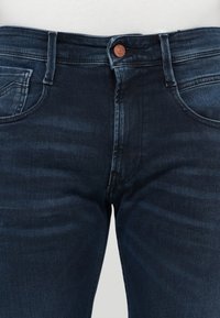 Jeans de denim azul oscuro con un diseño ajustado, que presentan un botón de cobre, bolsillos frontales y un ligero desteñido a lo largo de los muslos y las rodillas.
