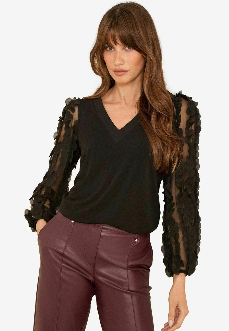 Top nero con scollatura a V e maniche a sbuffo nere trasparenti con un motivo floreale testurizzato e stratificato, abbinato a pantaloni bordeaux aderenti.