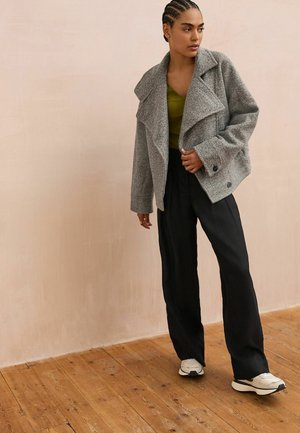 Grau melierte Wolljacke mit breiten Revers und doppelten Knöpfen, kombiniert mit einem grünen Oberteil und schwarzen weiten Hosen, vor einem hellen Hintergrund.