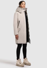 Vrouw in een beige knie-lange puffer jas met hoge kraag, zwarte leggings en beige robuuste veterschoenen, staand op een lichte achtergrond.