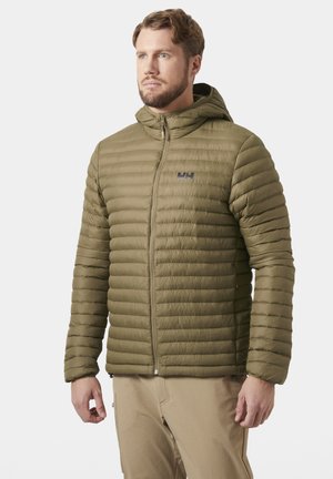 Hombre con chaqueta acolchada con capucha de color verde oliva y pantalones beige, de pie y mirando hacia un lado sobre fondo blanco.