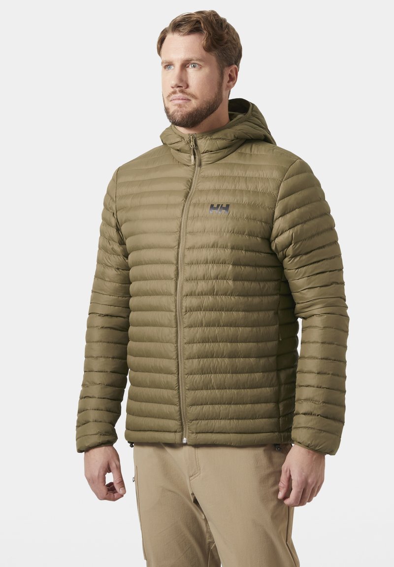 Hombre con chaqueta acolchada con capucha de color verde oliva y pantalones beige, de pie y mirando hacia un lado sobre fondo blanco.