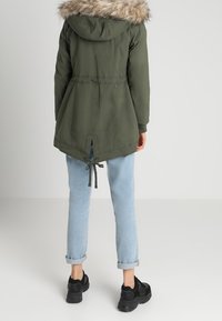 Parka verde oliva con capucha de piel sintética, cintura ceñida y cordón en el dobladillo, combinada con jeans azul claros remangados y zapatillas negras.