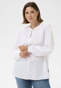 Blouse blanche avec encolure ronde, détail goutte d'eau et manches longues froncées. Confectionnée dans un tissu lisse, avec une coupe décontractée et des fentes latérales.