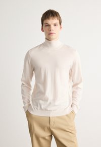 Pull en maille côtelée beige clair avec un col roulé, en tissu doux et lisse, à manches longues et ourlet côtelé. Associé à un pantalon beige.