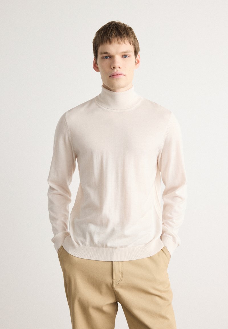 Pull en maille côtelée beige clair avec un col roulé, en tissu doux et lisse, à manches longues et ourlet côtelé. Associé à un pantalon beige.