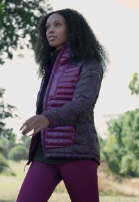 Chaqueta acolchada púrpura con un patrón geométrico, bolsillos con cremallera y un cuello alto. Llevada sobre una blusa negra y combinada con leggings color burdeos.