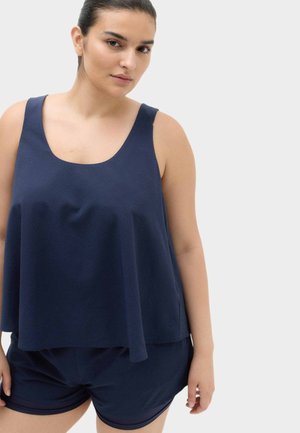 Kvinde med mørkt hår bundet tilbage ifører sig en løs mørkeblå ærmeløs top og matchende shorts, står mod en ensartet lys baggrund.