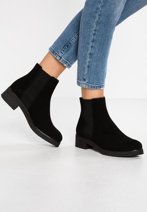 Sorte ruskind ankelstøvler med elastiske sidepaneler, en struktureret sort sål og en lav blokhæll, placeret mod en hvid baggrund.