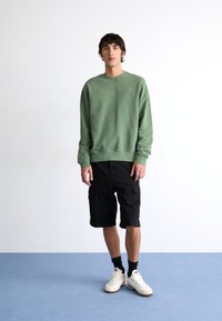Green sweatshirt with ribbed cuffs and hem, paired with black cargo shorts. Model wears white sneakers and black socks, standing on blue carpet.